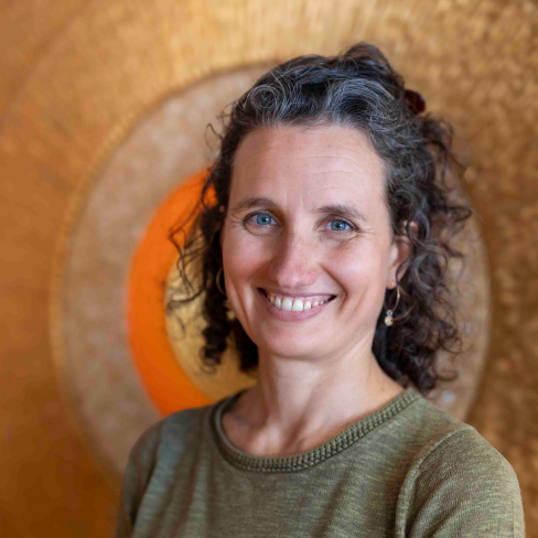 Ein lächelnde Frau mit lockigem Haar steht vor einem grossen Sonnenmandala.